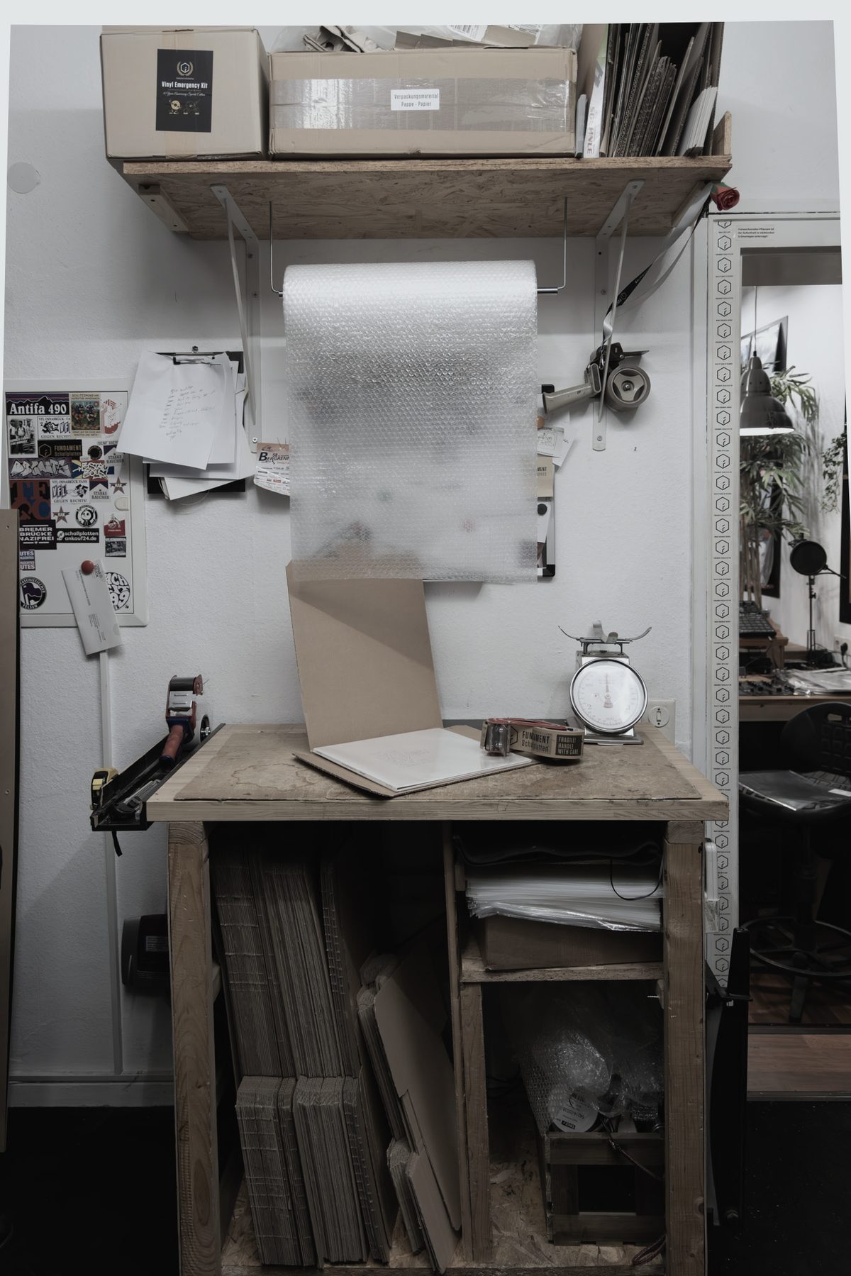 Die Packstation im Laden: Arbeitstisch mit Waage, Rolle Verpackungsmaterial, Kartons und Versandtaschen im Regal darunter.